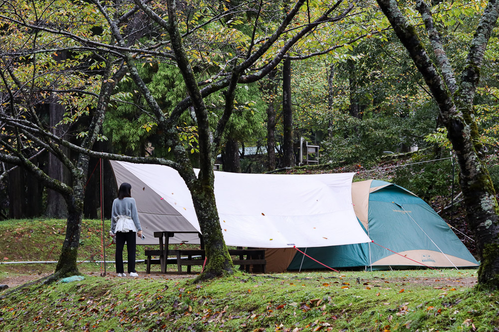 A young women standing in front of a large camping area, with a tent surrounded by trees on a rainy day.