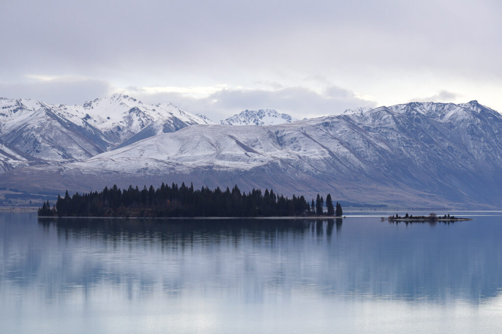 The view of Motuariki in Lake Tekapo with snowy mountain at the back, and a slightly reflective lake water.