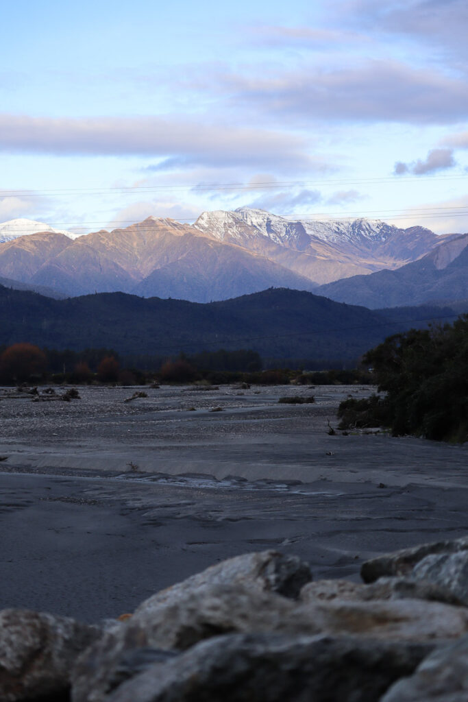 Southern Alps from Hokitika River, South Island NewZealand, with the sun shining on the alps and casting shadow on the river bank
