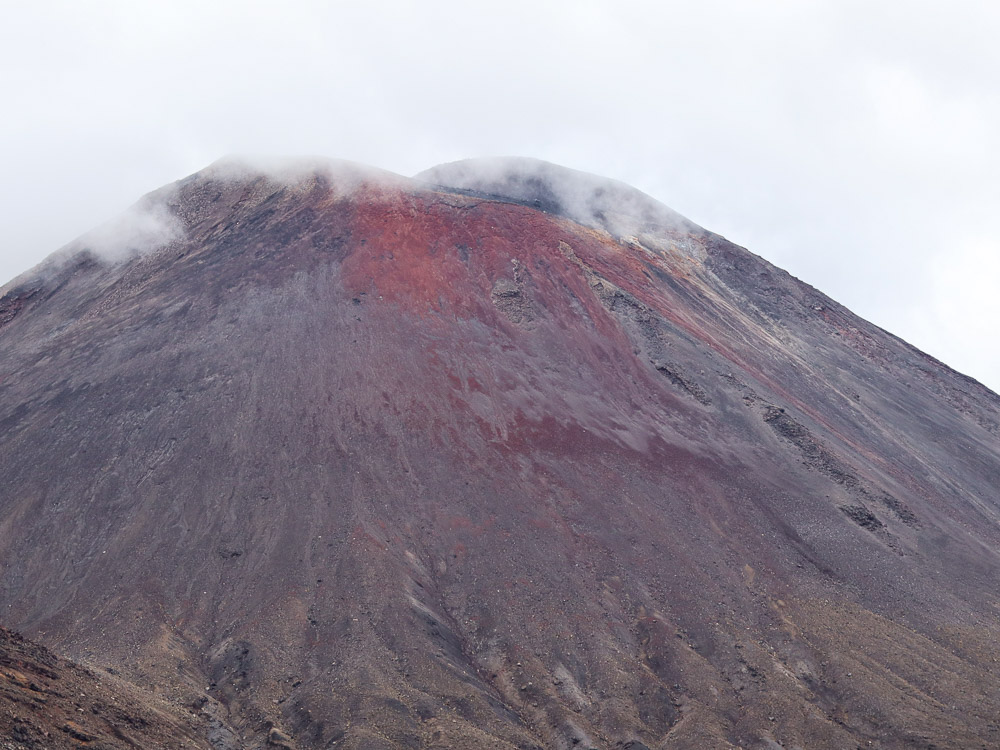 The peak of Mount Ngauruhoe from The Lords of The Rings, covered in clouds with the top slightly visible and the rusty red scoria popping out.