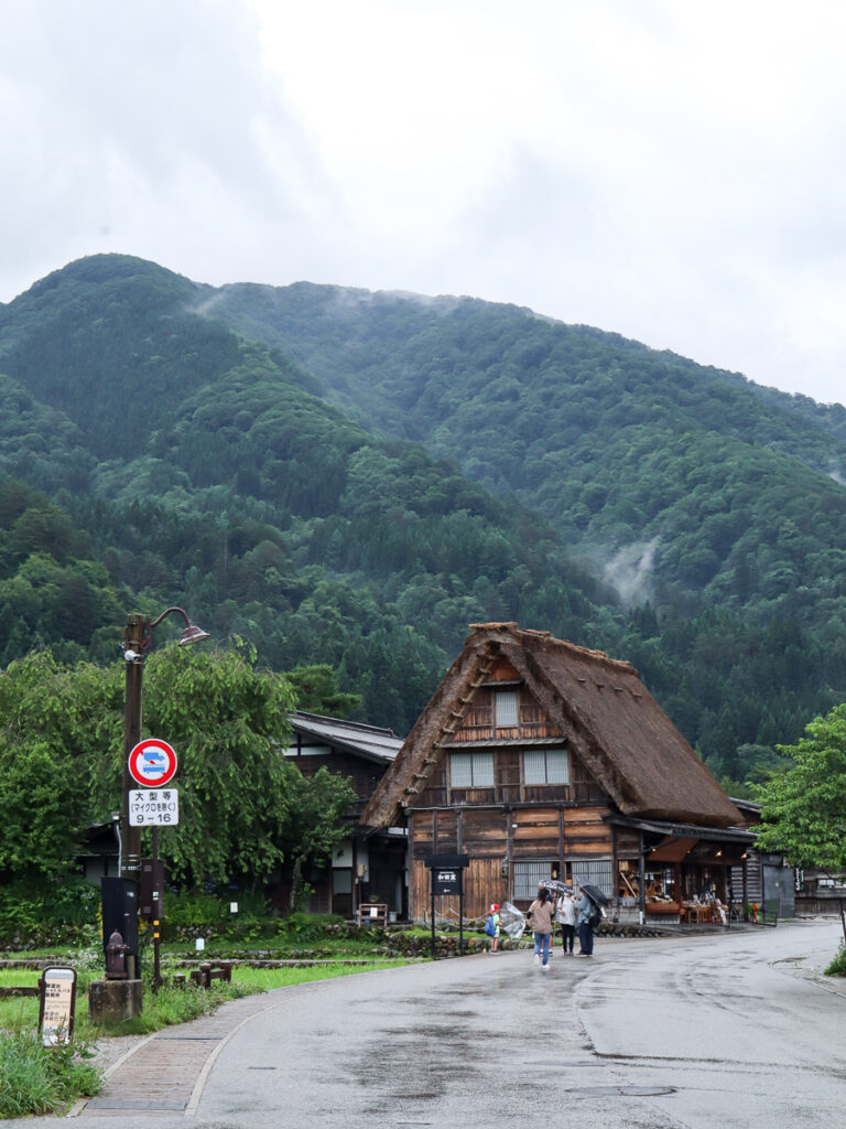 Shirakawa-go in summer, a light rainfall making the road and the historical house slightly wet, and people holding on to their umbrellas.