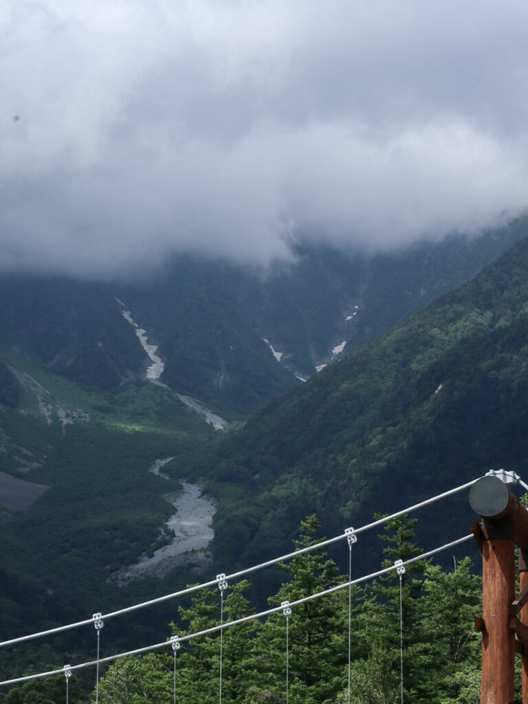 Kappa Bridge and Azusa river on the Hotaka mountain range behind. The scale of the mountain towers over the scenery, its tall peak covered with dark clouds.