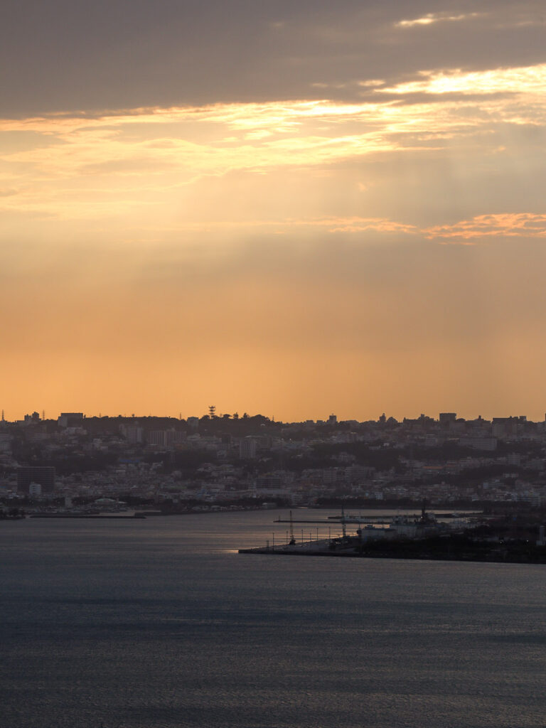 Sunset in Okinawa. The sky dyed orange, and rays of light pierces through the clouds onto the sea and the city.