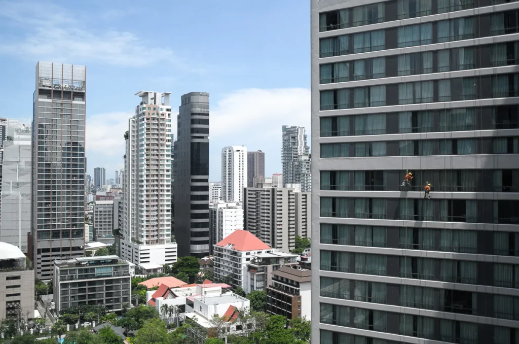 The view from a hotel room out to the Benchasiri park on the left with other buildings of bangkok city. The right side is the opposite guest quarter building with two glass cleaners hanging down.