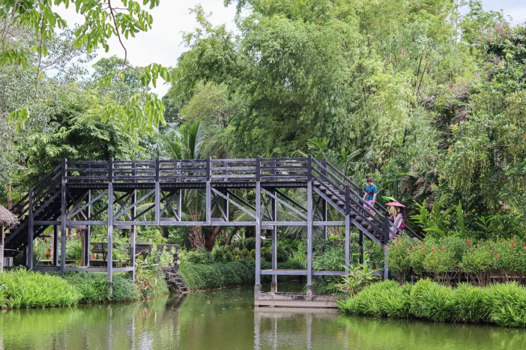 A tall wooden bridge at Mallika R.E. 124 in Kanchanaburi, with people in Thai traditional dress.